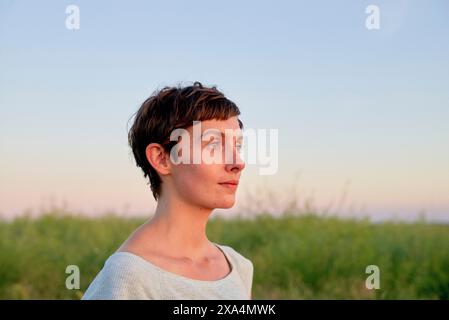 Une jeune femme aux cheveux courts se tient dans un champ au crépuscule, regardant sur le côté avec une expression calme. Banque D'Images