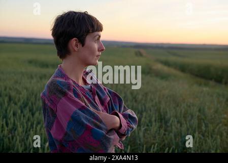 Une jeune femme se tient dans un champ au crépuscule, les bras croisés tout en regardant au loin avec une expression sereine. Banque D'Images