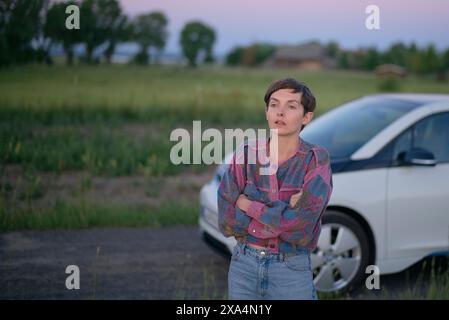 Une jeune femme aux cheveux courts se tient près d'une voiture devant une scène de campagne au crépuscule, paraissant contemplative. Banque D'Images