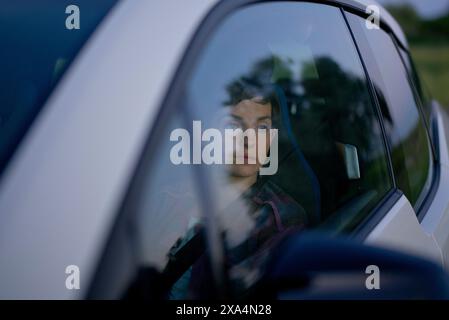 Une jeune femme regarde à l'extérieur depuis le siège passager d'un véhicule, avec leur reflet visible dans la fenêtre de la voiture. Banque D'Images