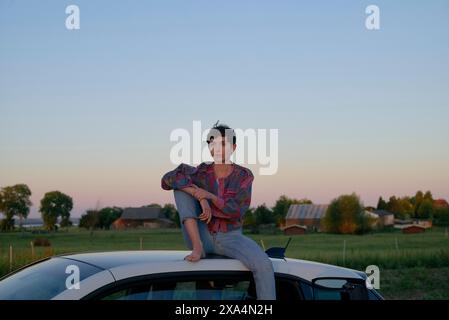 Une jeune femme est assise réfléchie au sommet d'un toit de voiture au crépuscule, avec des arbres et des bâtiments dans le paysage rural lointain. Banque D'Images