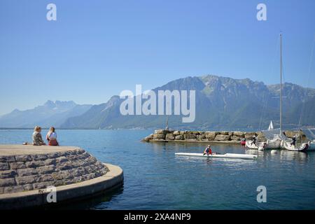 Une vue panoramique sur le lac où deux personnes sont assises sur le bord d'une jetée, regardant l'eau où une personne fait du kayak près de voiliers amarrés, avec des montagnes en arrière-plan sous un ciel bleu clair. Banque D'Images