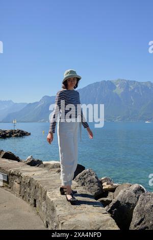 Une personne se tient debout sur un rivage rocheux avec un grand plan d'eau et des montagnes en arrière-plan, portant un chapeau, un haut rayé et un pantalon blanc. Banque D'Images