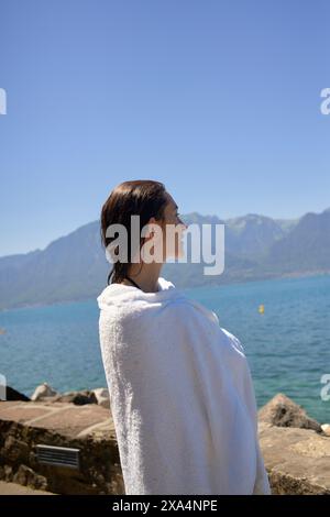 Une personne enveloppée dans une serviette blanche se tient près d'un lac regardant les montagnes sous un ciel bleu clair. Banque D'Images