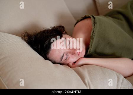 Une jeune femme dort paisiblement sur un canapé beige, portant un dessus vert, la tête reposant sur un coussin blanc. Banque D'Images