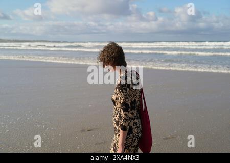 Une jeune femme aux cheveux aux épaules, vêtue d'une robe et portant un sac rouge, se tient sur une plage de sable avec des vagues en arrière-plan. Banque D'Images