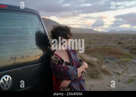 Une femme s’appuie contre un véhicule Volkswagen avec un pare-brise fissuré, regardant au loin un paysage désolant sous un ciel nuageux de coucher de soleil. Banque D'Images