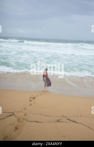Une jeune femme se tient sur une plage de sable, regardant les vagues avec des empreintes de pas derrière elles. Banque D'Images