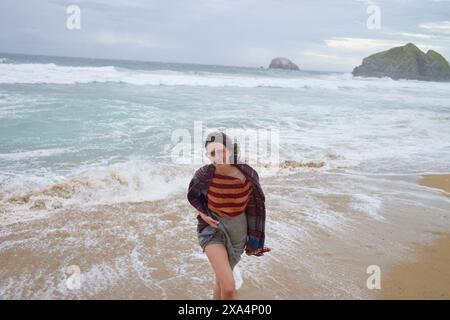 Une femme sourit en courant sur une plage de sable avec des vagues et deux gros rochers visibles en arrière-plan sous un ciel nuageux. Banque D'Images