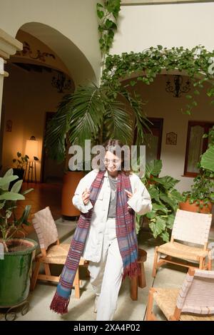 Une jeune femme souriante marche dans un espace intérieur avec des plantes en pot, tenant une écharpe colorée et portant des vêtements décontractés et élégants. Banque D'Images