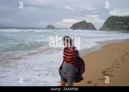 Une femme marche le long d'une plage de sable avec des vagues qui s'écrasent en arrière-plan, portant un pull rayé et tenant sa jupe contre le vent. Banque D'Images