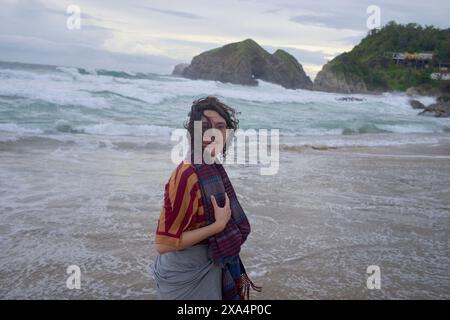 Une personne se tient debout sur une plage de sable avec des vagues qui s'écrasent à terre, face à la caméra avec un sourire, et portant un haut rayé et une écharpe, avec un affleurement rocheux et un ciel nuageux en arrière-plan. Banque D'Images