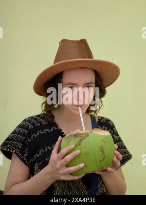 Une jeune femme portant un chapeau brun sirote d'une noix de coco avec une paille sur un fond jaune pâle. Banque D'Images