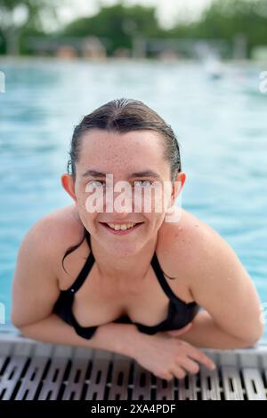 Une jeune femme souriante aux cheveux mouillés est dans une piscine, se reposant sur le bord de la piscine et regardant directement la caméra. Banque D'Images