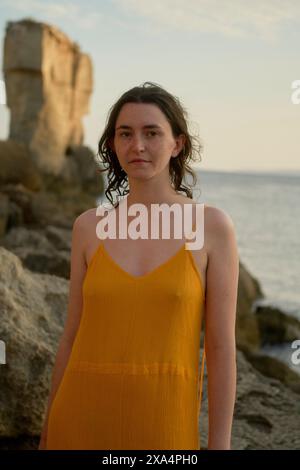 Une femme en robe jaune se tient devant une côte rocheuse pendant le coucher du soleil. Banque D'Images