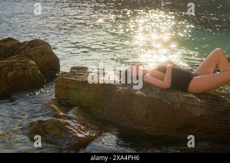 Une jeune femme se prélasse sur un rocher au bord de la mer, trempant au soleil avec ses mains reposant sur leur section médiane. Banque D'Images