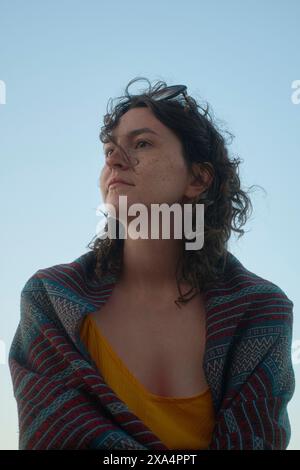 Une jeune femme pensive avec des cheveux bouclés et des lunettes sur la tête regarde au loin, drapée dans un châle à motifs contre un ciel clair. Banque D'Images