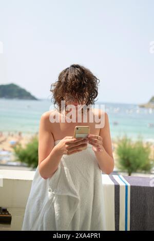 Une femme enveloppée dans une serviette se tient sur un balcon à l'aide d'un smartphone, avec une plage et la mer en arrière-plan. Banque D'Images