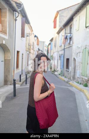 Une jeune femme aux cheveux aux épaules sourit et regarde par-dessus son épaule tout en marchant dans une rue étroite bordée de maisons traditionnelles de style européen. Banque D'Images