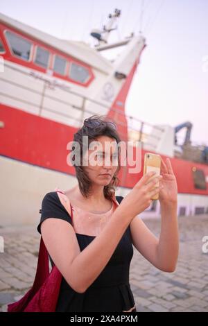 Une femme prend un selfie avec un smartphone devant un bateau rouge et blanc. Banque D'Images