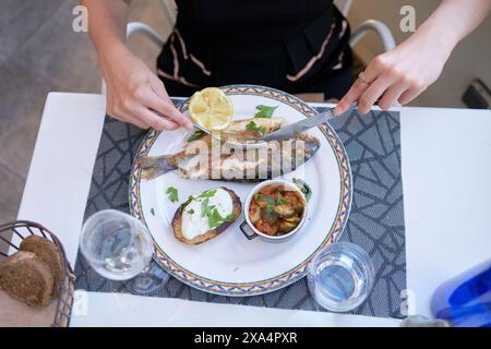 Une personne est assise à une table, sur le point de profiter d'un repas composé d'un poisson grillé garni de citron, d'un côté de pain, d'une sauce crémeuse, et d'un petit bol de ce qui ressemble à des légumes cuits. Banque D'Images