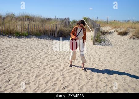 Une jeune femme marche sur un sentier de plage de sable à côté de clôtures en bois, sous un ciel bleu clair. Banque D'Images