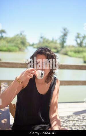 Une personne est assise à l'extérieur à une table en bois sirotant une tasse de café avec un fond d'eau pittoresque et un temps ensoleillé. Banque D'Images
