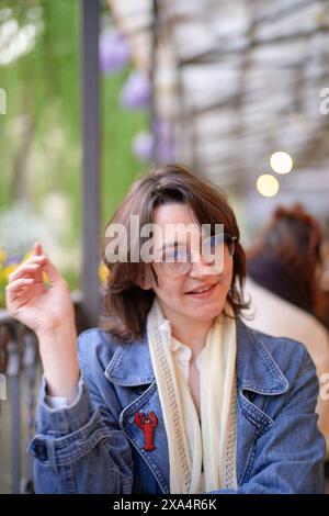 Une femme souriante est assise dans un café en plein air, tenant une fleur jaune, portant une veste en denim, des lunettes et une écharpe blanche. Banque D'Images