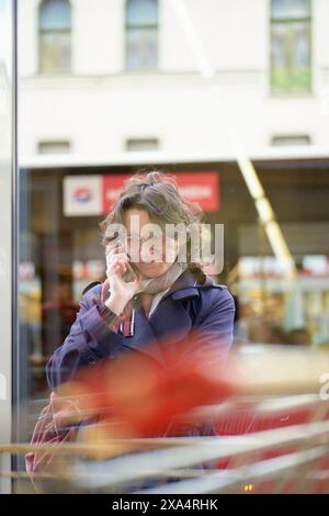 Femme en manteau bleu parlant au téléphone derrière un objet rouge flou dans la ville. Banque D'Images