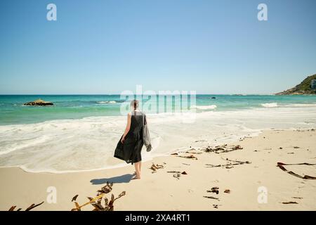 Femme marchant seule sur une plage ensoleillée avec des vagues en arrière-plan et des algues éparpillées sur le sable. Banque D'Images