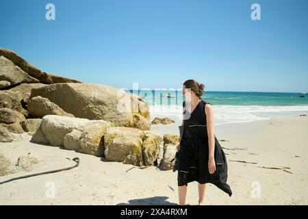 Femme marchant sur une plage ensoleillée avec de grands rochers en arrière-plan. Banque D'Images
