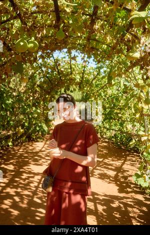 Femme souriante en robe rouge debout sous une pergola de vigne verte écrivant dans un cahier avec la lumière du soleil filtrant à travers les feuilles. Banque D'Images