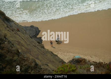 Deux personnes marchant sur une plage de sable à côté d'une falaise avec des vagues se brisant en arrière-plan. Banque D'Images