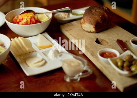 Un petit-déjeuner sain est servi sur une table en bois avec un bol de fruits hachés, des tranches de fromage, une miche de pain et un assortiment de condiments. Banque D'Images