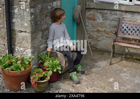 Un enfant est assis à côté de plantes en pot sur une marche de pierre près d'un banc en bois rustique et une serpillière appuyée contre un vieux mur. Banque D'Images