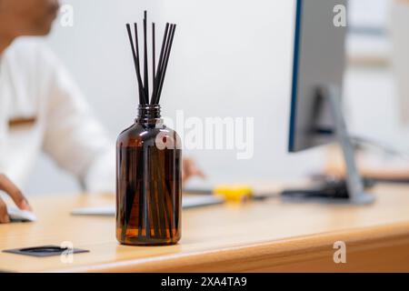 Une bouteille en verre brun avec des bâtons de roseau noirs sur un bureau en bois dans un cadre de bureau. Une personne travaille sur un ordinateur en arrière-plan. Banque D'Images