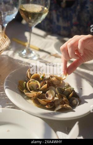 Gros plan d'une main cueillant une palourde dans une assiette de palourdes cuites à la vapeur, avec un verre de vin blanc en arrière-plan. Banque D'Images