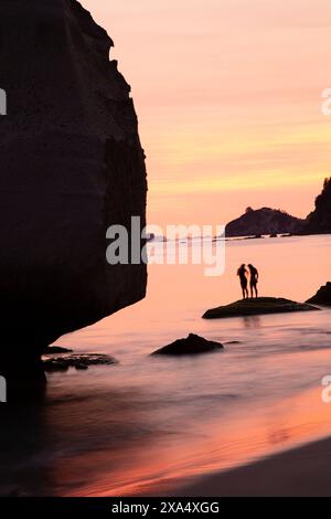 Couple silhouetté sur une plage au coucher du soleil avec des formations rocheuses dures et réflexion sur l'eau. Banque D'Images