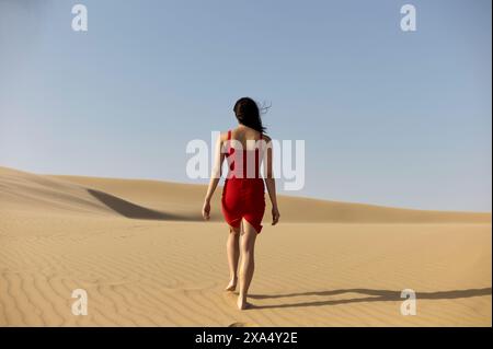 Femme dans une robe rouge marchant sur une dune de sable sous un ciel clair. Banque D'Images