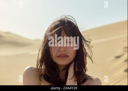 Femme embrassée par le soleil avec les yeux fermés et les cheveux tourbillonnés par le vent se tient au milieu du sable doré du désert. Banque D'Images