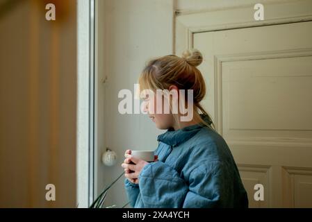 Femme dans un manteau matelassé bleu tenant une tasse blanche et regardant soigneusement par une fenêtre. Banque D'Images