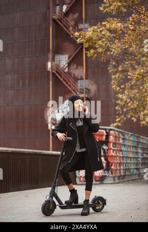 Jeune femme parlant sur son smartphone tout en se tenant debout avec un scooter électrique un jour d'automne dans une rue de la ville .. Banque D'Images