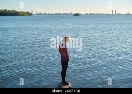 Femme debout sur un rocher au bord d'un lac calme regardant vers la ligne d'horizon au loin pendant la lumière du jour. Banque D'Images