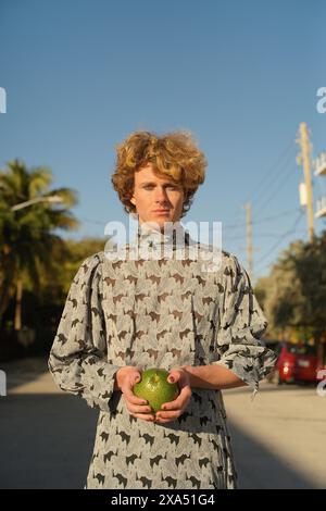 Jeune homme aux cheveux bouclés debout à l'extérieur tenant une pomme verte, portant une chemise à motifs avec des arbres. Banque D'Images