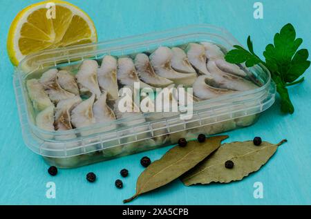 Poisson mariné dans un récipient en plastique. Morceaux de hareng mariné dans un emballage ouvert d'un magasin sur un fond turquoise. Banque D'Images