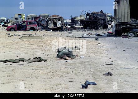 Première Guerre du Golfe : 4 mars 1991 le corps d'un soldat repose dans le désert avec un assortiment de véhicules militaires et civils détruits d'un convoi militaire irakien formant une toile de fond. Il a été attaqué sur la route 801, la route menant à Um Qasr, au nord de Koweït City, par l'armée de l'air américaine alors que l'armée irakienne tentait de fuir vers l'Irak. Banque D'Images