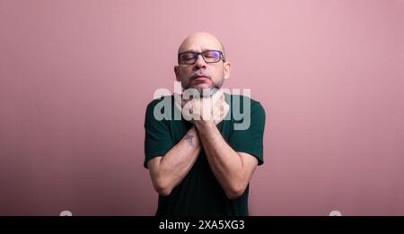 Homme chauve barbu avec des lunettes de vue serrant son cou avec les deux mains. Isolé sur fond de couleur saumon. Banque D'Images