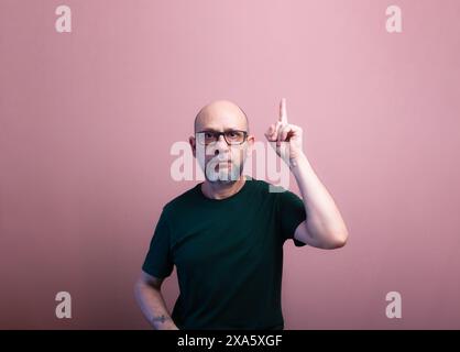 Homme chauve barbu avec des lunettes de vue pointant vers le haut avec son doigt. Isolé sur fond de couleur saumon. Banque D'Images