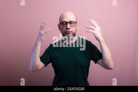 Portrait d'homme chauve et barbu avec des lunettes avec les deux mains à côté de sa tête avec les yeux fermés. Isolé sur fond de couleur saumon. Banque D'Images