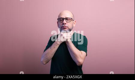 Homme chauve barbu avec des lunettes de vue serrant son cou avec les deux mains. Isolé sur fond de couleur saumon. Banque D'Images